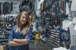 Mulher sorrindo em uma distribuidora automotiva, com os braços cruzados, cercada por ferramentas e peças organizadas na parede ao fundo.